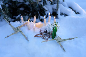 altar for Imbolc sabbath, pagan holiday ritual. Brigid's cross of straw, candles, snowdrops, toy sheep on snow, winter forest natural background. symbol of Imbolc holiday,